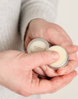 A close-up of a person's hands holding small silver metal tins of Nail & cuticle balm, with one tin open to show a pale cream-colored balm. The person is wearing a light gray knit sweater against a plain, light background.