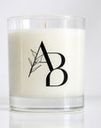A white wax candle in a clear glass jar with a black monogram logo featuring a leaf design, set against a clean white background.
