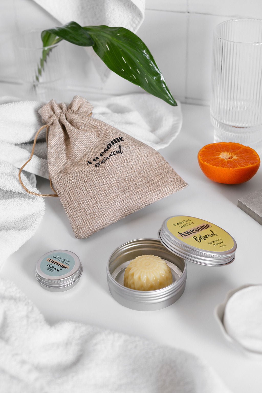 Hessian Awesome Botanical Bag in bathroom with Lip Balm & Hand Balm surrounded by a plant and fresh orange 