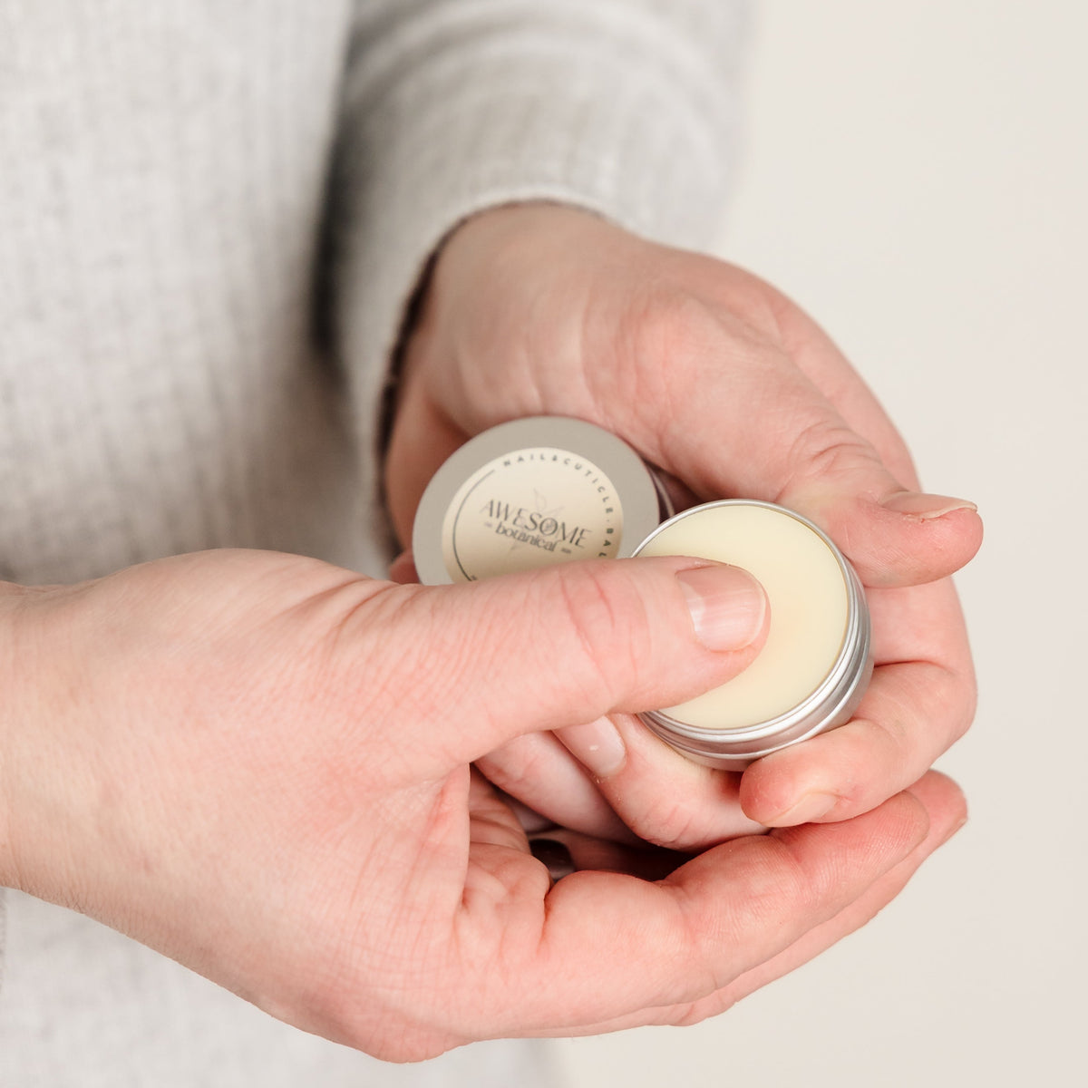 A close-up of a person's hands holding small silver metal tins of Nail &amp; cuticle  balm, with one tin open to show a pale cream-colored balm. The person is wearing a light gray knit sweater against a plain, light background.