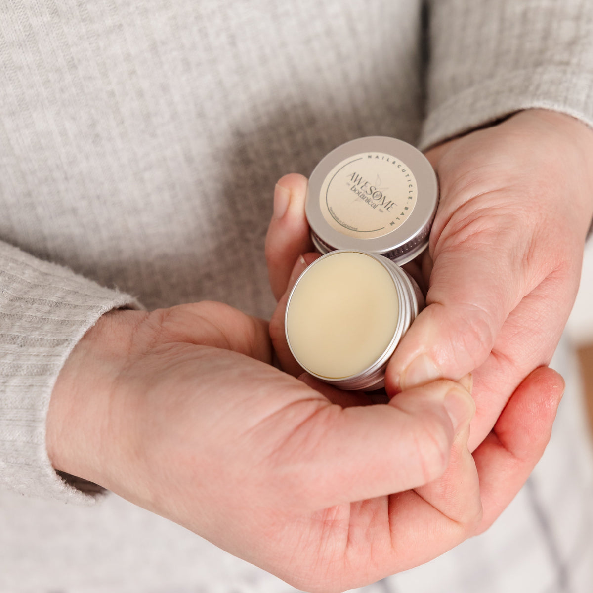 A close-up of a person's hands holding  small silver tins of nail &amp; cuticle balm. One tin is open, showing a pale yellow balm, and the other is closed with a label that reads Nail &amp; cuticle Balm