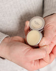 A close-up of a person's hands holding  small silver tins of nail & cuticle balm. One tin is open, showing a pale yellow balm, and the other is closed with a label that reads Nail & cuticle Balm