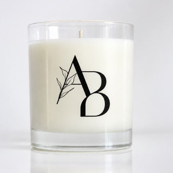 A white wax candle in a clear glass jar with a black monogram logo featuring a leaf design, set against a clean white background.