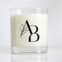 A white wax candle in a clear glass jar with a black monogram logo featuring a leaf design, set against a clean white background.